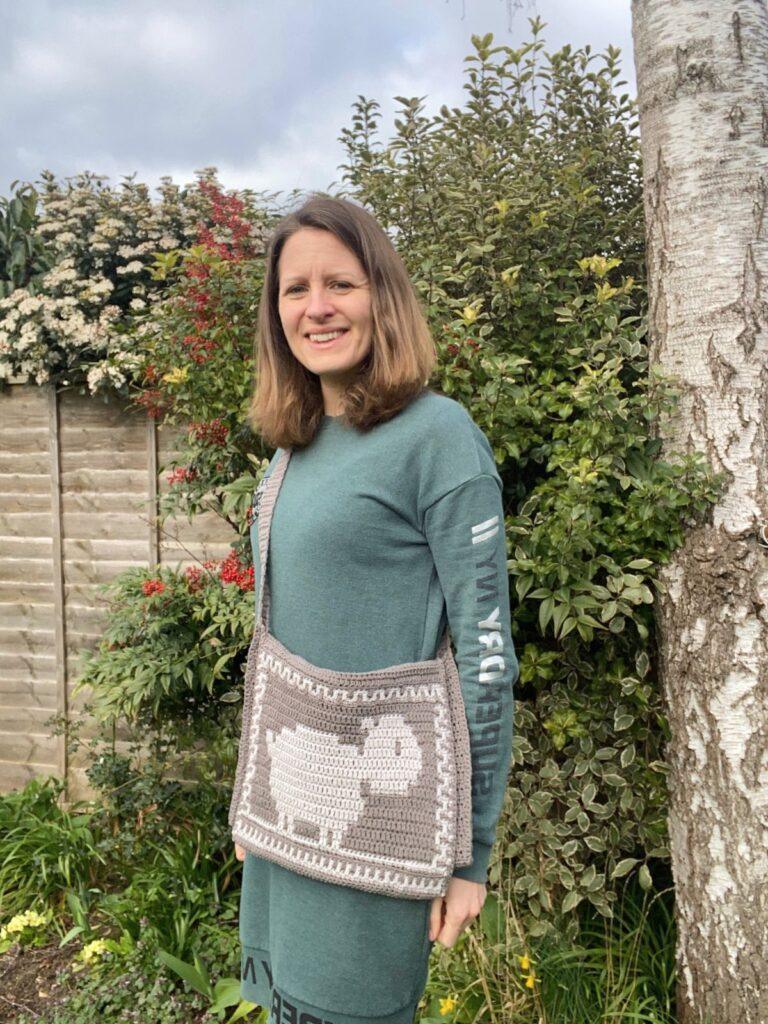A lady stands in a garden wearing a green sweater and a grey crochet shoulder bag with a sheep on the front