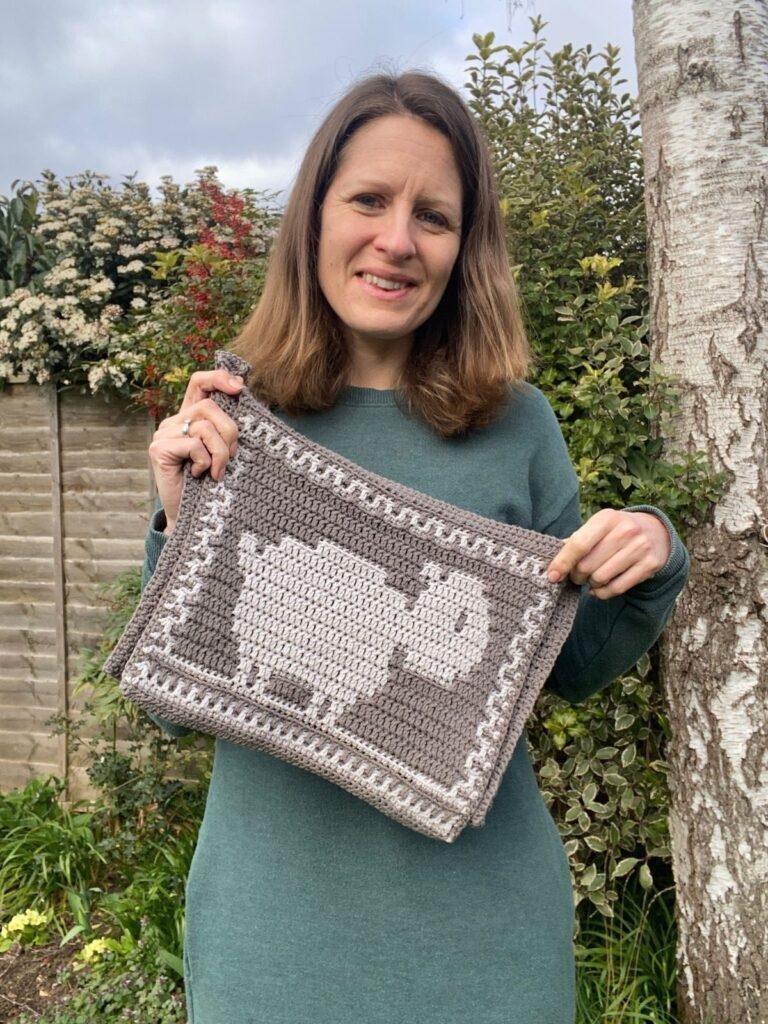 A person in a green jumper dress hold up a grey and white crochet bag with a sheep on the front