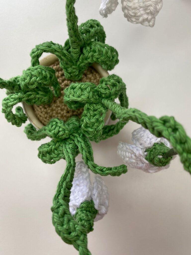 A top down view of a pot of crochet flowers with lots of green leaves showing and white flowers