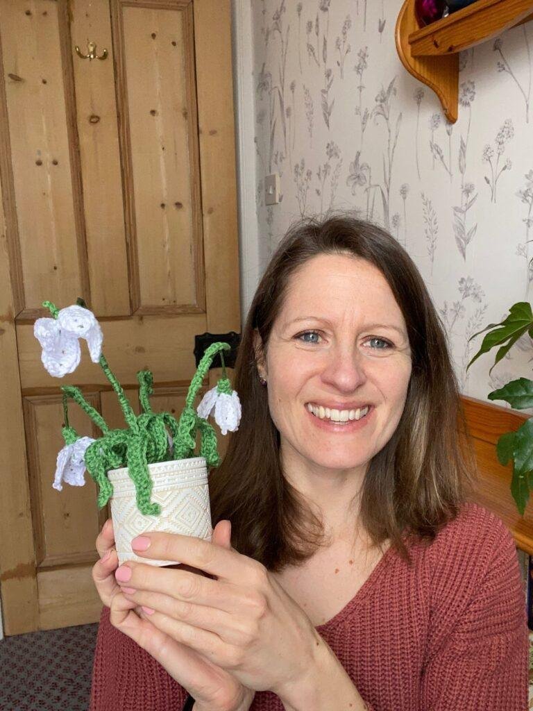 A woman smiles and holds a crochet snowdrop in a pot with a wooden door behind her