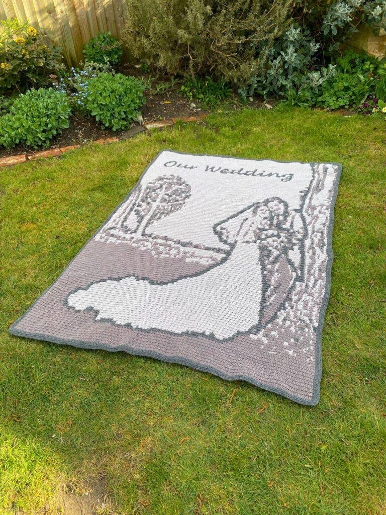A large crochet blanket lies outside on a lawn. The blanket is grey with a bride and groom on it