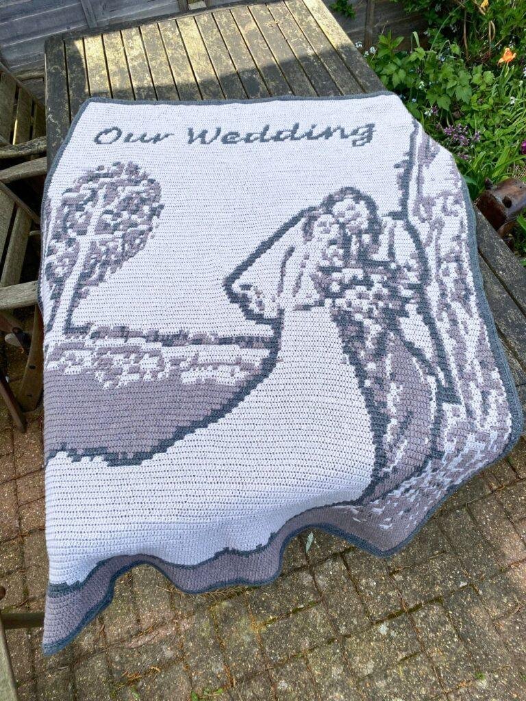 A crochet wedding blanket is laid over a table in a garden