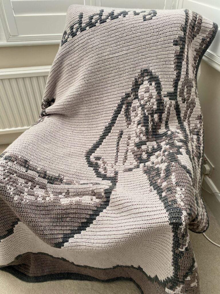 A crochet wedding themed blanket is draped over a chair by a radiator