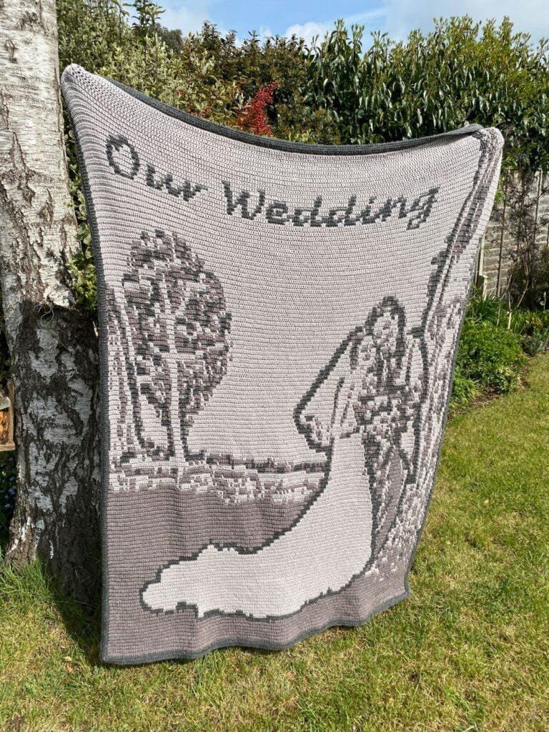 A grey crochet blanket is held up in a garden, hiding the person behind it. It features a bridal couple and the words our wedding