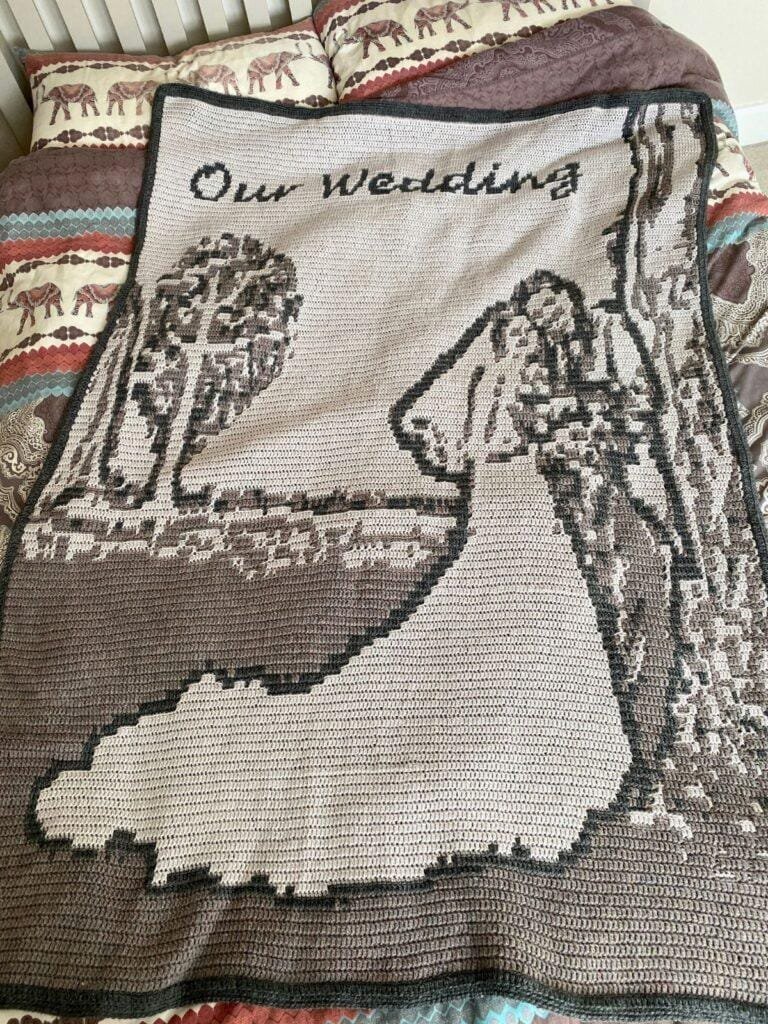 A crocheted blanket featuring a married couple and the words our wedding, is laid on a bed with a patterned duvet