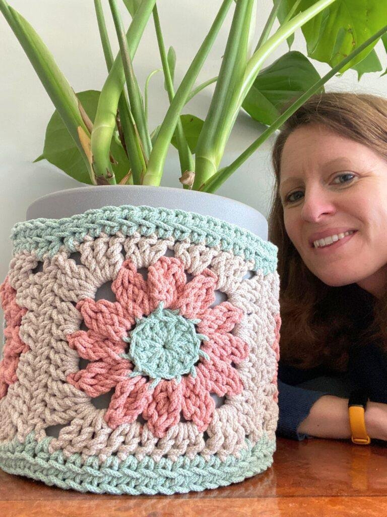 A lady peers into the photo next to a large crochet plant pot cover over a plant pot holding a plant