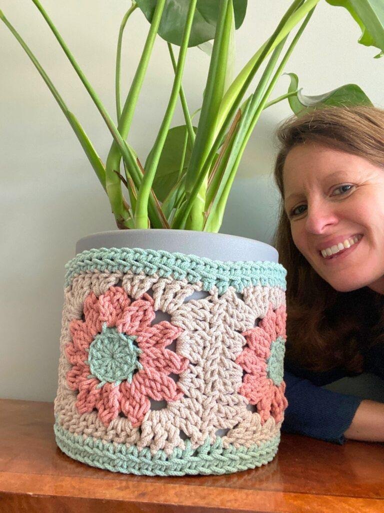 A large plant with a crochet plant pot cover sits on a shelf and a person leans into the shot smiling