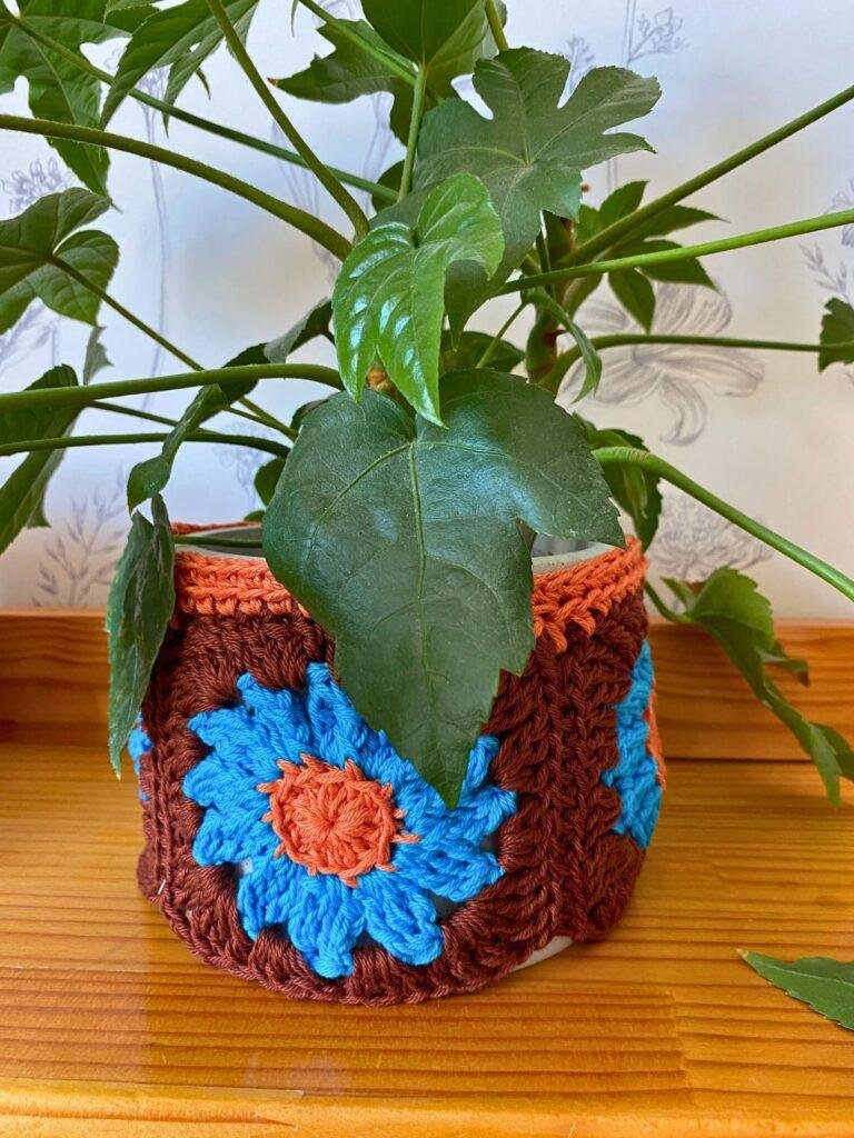 A green plant is in a pot covered with a brown, orange and blue crochet plant pot cozy.