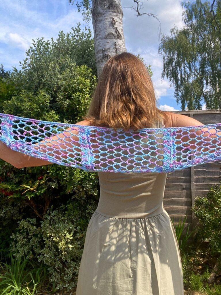 A lady stands with her back to the camera in a garden holding a blue crochet lacy scarf out across her back