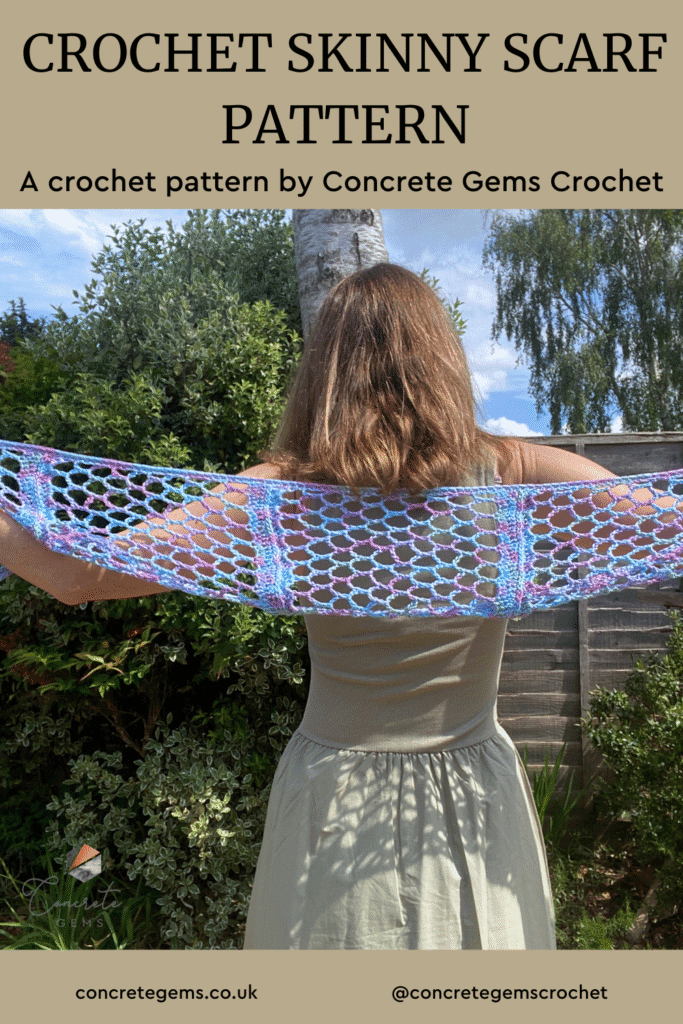 A crochet skinny scarf is being held out across a lady's back in a garden