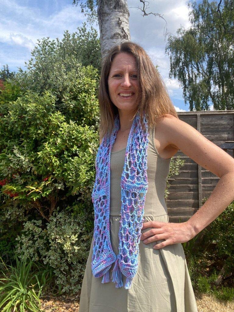 a lady stands in a garden with her hands on her hips wearing a green dress and a blue crochet lightweight scarf