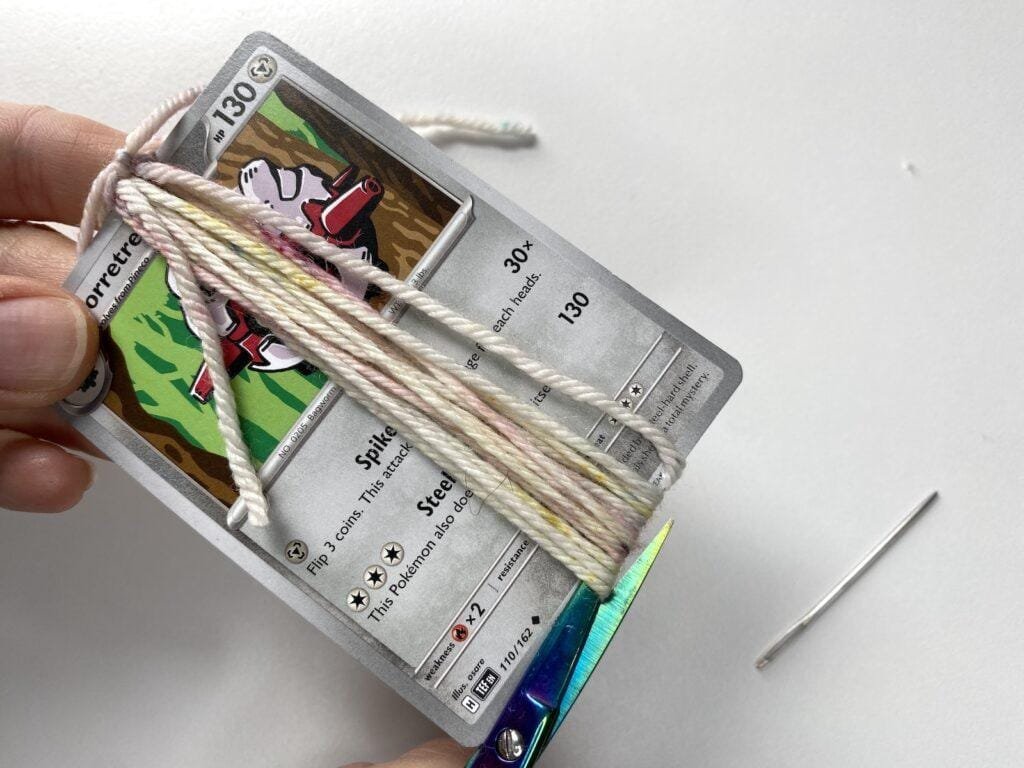 A hand is cutting some yarn wrapped round a piece of card with holographic scissors