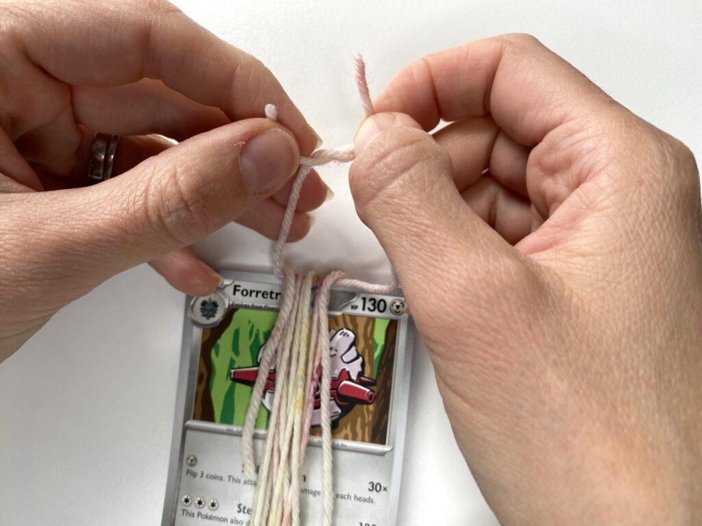 2 hands tying a piece of yarn around some yarn which is wrapped around a card