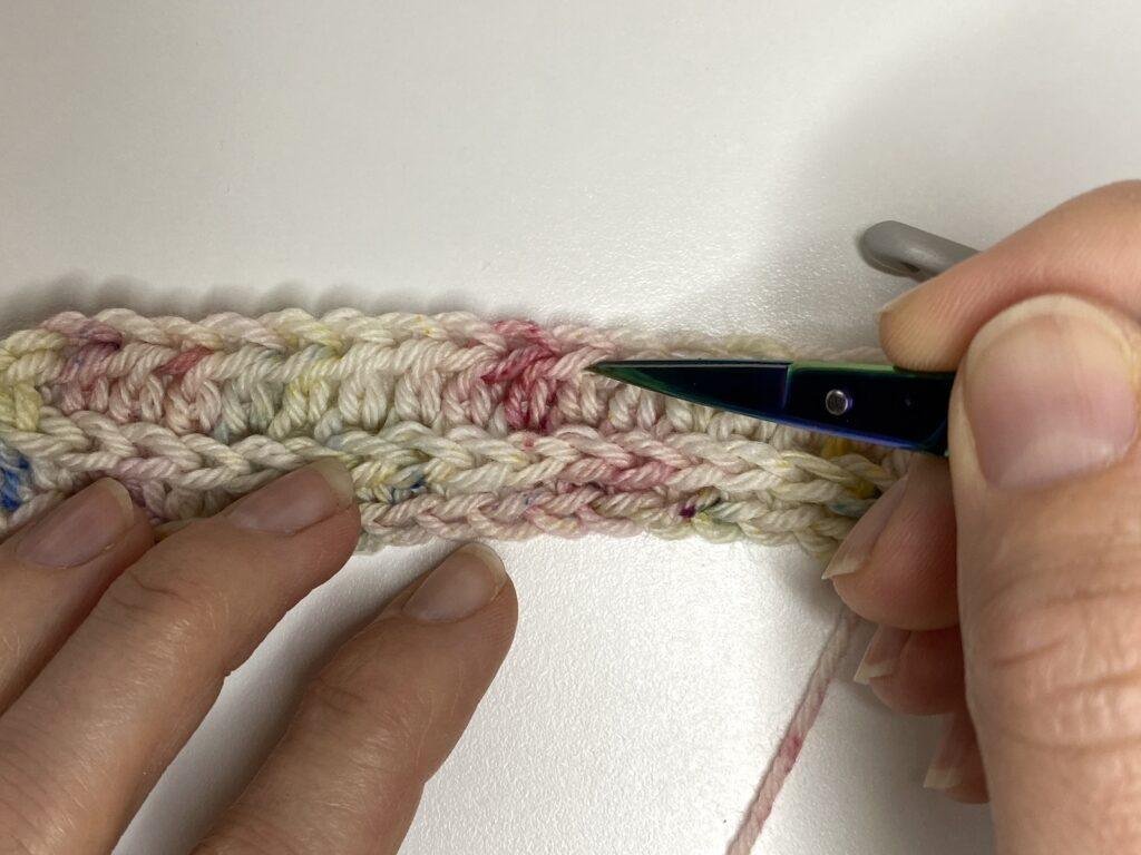 A person points at the third loop of a row of half double crochet using a pair of scissors