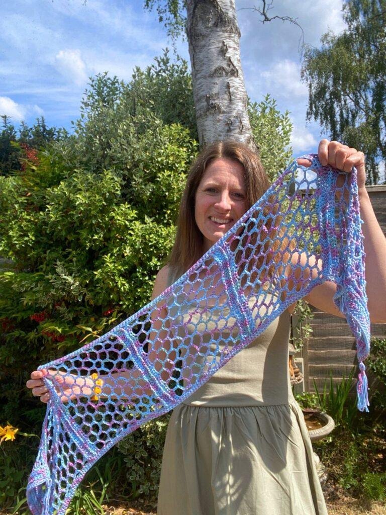 A lady stands in a garden holding out a lacy crochet scarf in blues and pinks