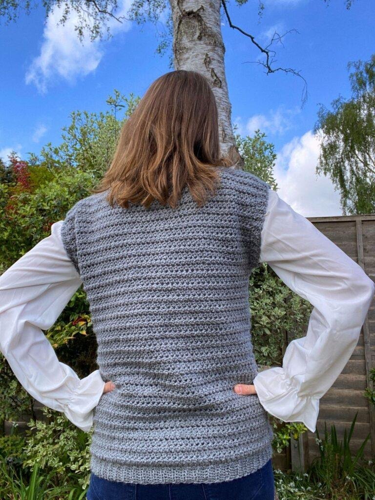 A lady with her back to the camera in a garden wears a long sleeved white shirt with a grey crochet tank top over the top
