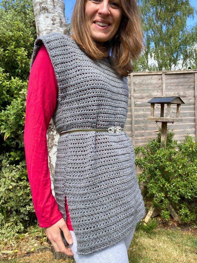 A woman in a garden is wearing a crochet grey tunic over a pink long sleeve top, with a silver belt