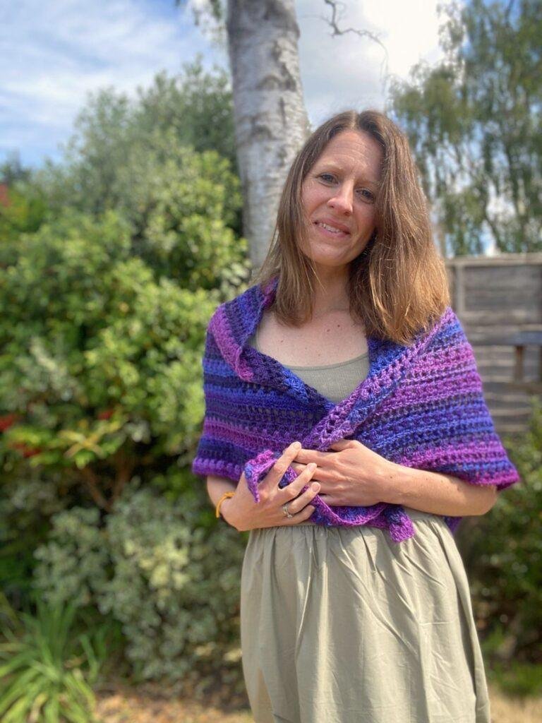 A lady wears a green dress and holds a purple crochet shawl around her shoulders. She is standing by a tree and bush with her head on one side