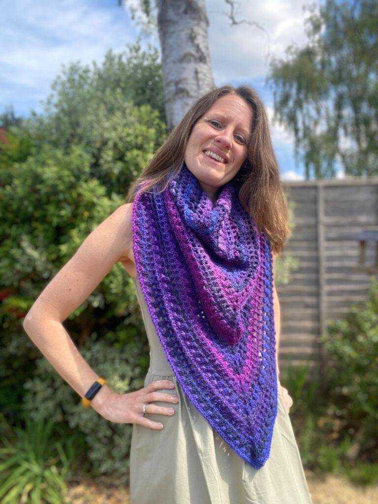 A smiling lady with her hands on her hips in the garden, is wearing a green dress and a large purple crochet triangle granny stitch shawl round her neck