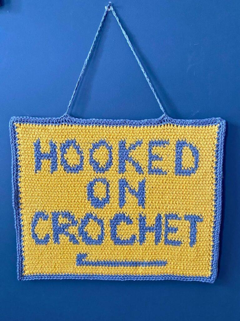 A yellow and grey crochet wallhanging featuring graphic words 'hooked on crochet' hang on a very dark grey wall