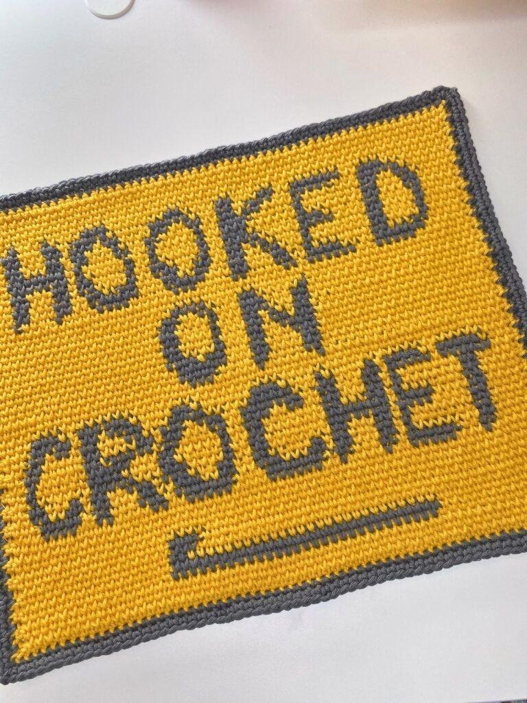 A yellow and grey crochet rectangle is slanted in the photo and features the words hooked on crochet which have been crocheted on using the waistcoat stitch