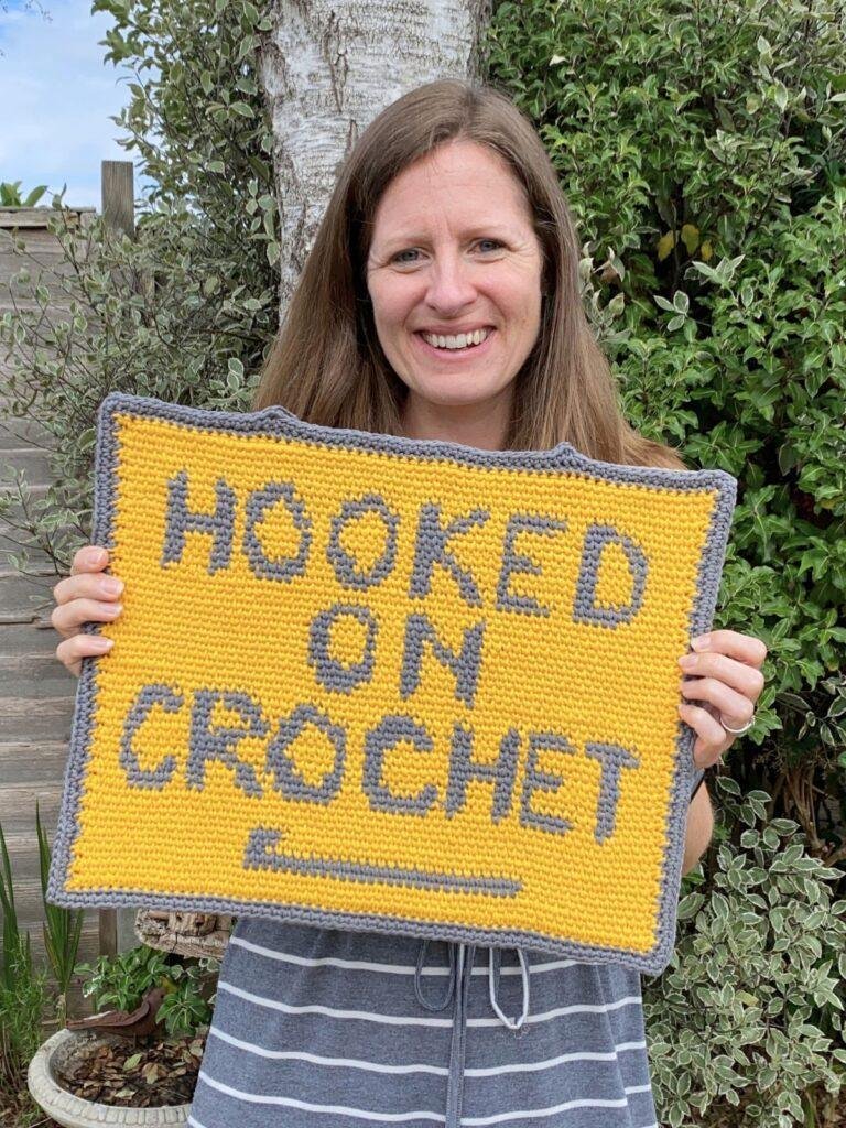 A person stands in a garden holding up a rectangular crochet picture featuring the words Hooked on Crochet