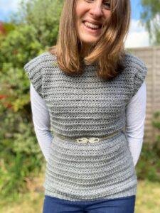 A lady with her arms behind her back wears a grey crochet sleeveless top and a silver belt over a white long sleeve top