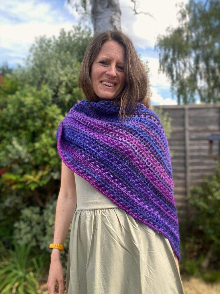 A lady is wrapped up in a crochet triangle shawl standing in a garden and smiling