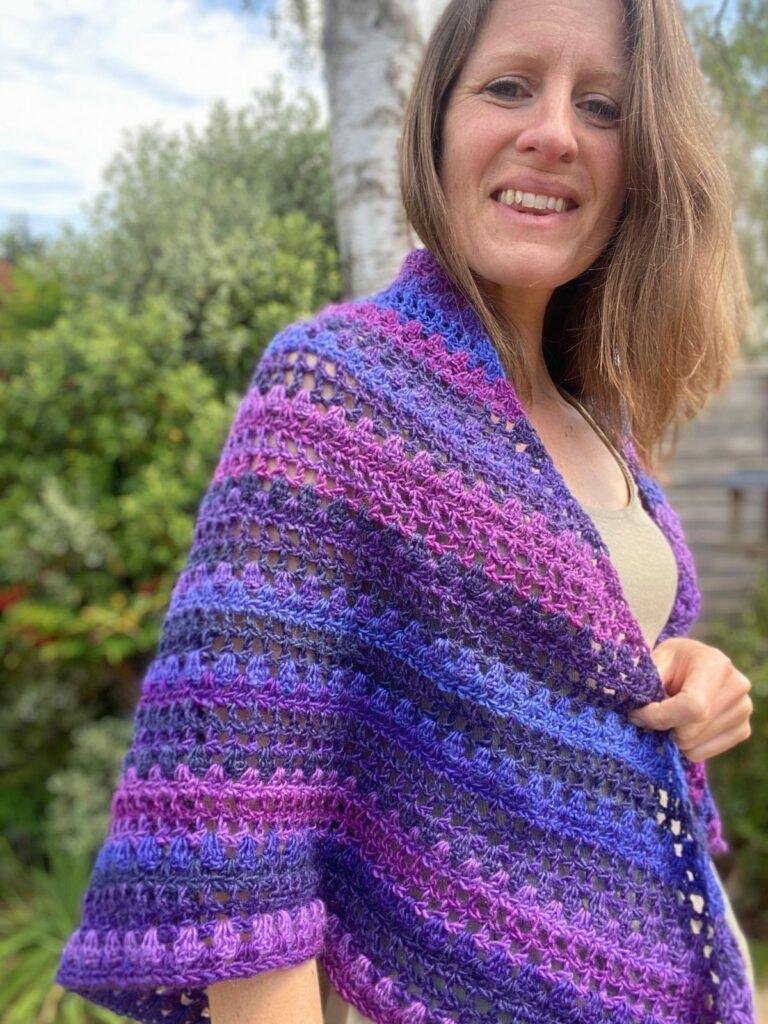 A lady stands in a garden with a purple crochet triangle shawl wrapped round her