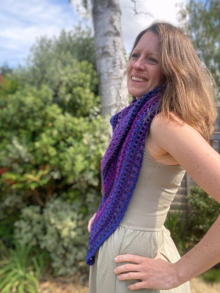 A woman with her hands on her hips leaning back to the side wearing a green sleeveless dress with a purple crochet shawl