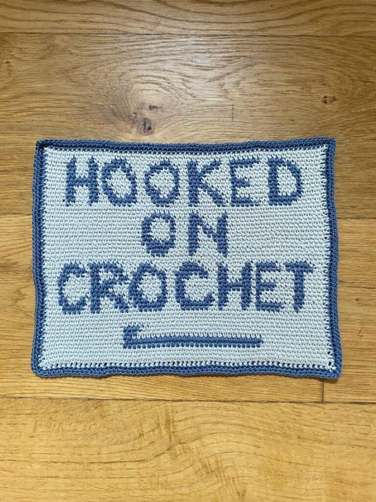A crochet wall hanging hangs on a wooden wall featuring the words hooked on crochet in blue with a beige background