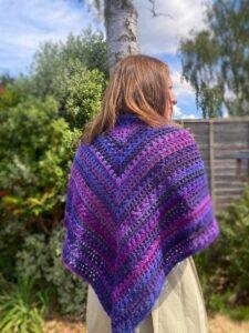 A lady stands with her back to the camera wearing a purple crochet triangle shawl