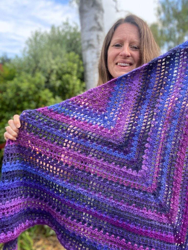 A lady holds up a variegated purple triangle crochet shawl, as she smiles with a tree and greenery in the background