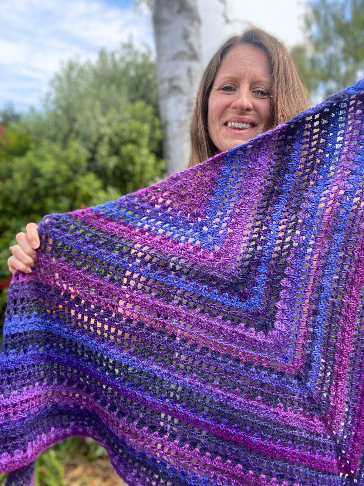 A lady holds up a variegated purple triangle crochet shawl, as she smiles with a tree and greenery in the background