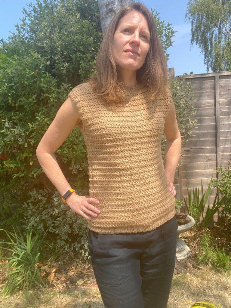 A lady stands in a sunny garden wearing a crochet tank top and black trousers