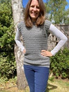 A lady is wearing jeans, a long sleeved white top and a grey crochet tank top over the top, standing in front of a tree