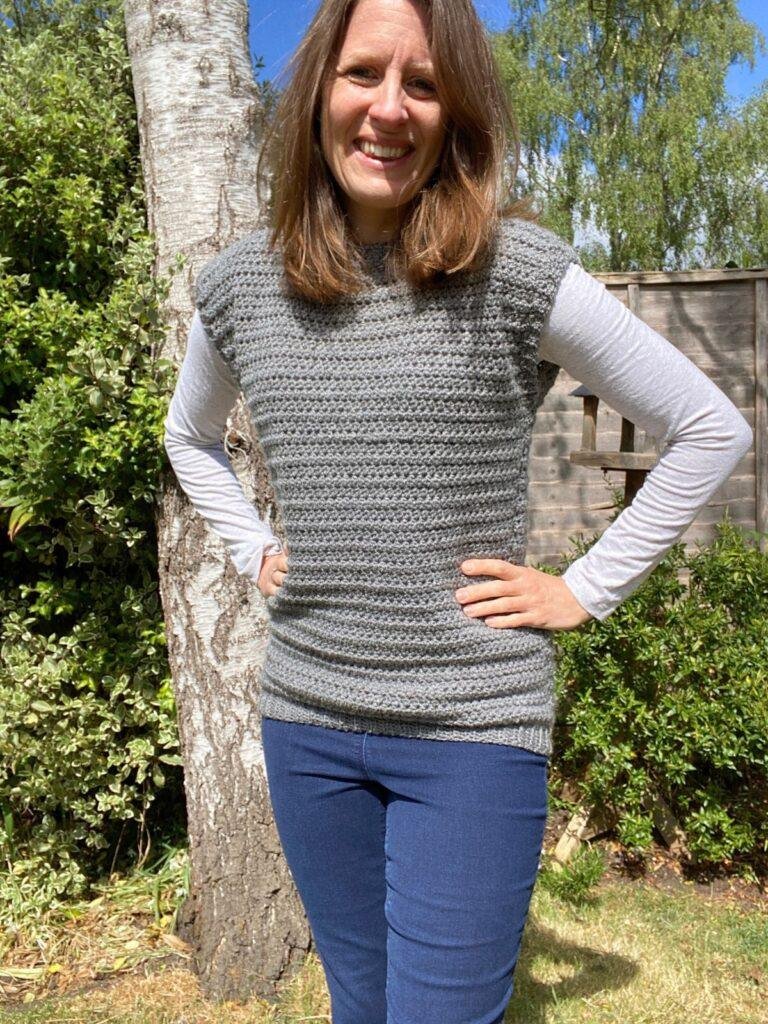 A lady is wearing jeans, a long sleeved white top and a grey crochet tank top over the top, standing in front of a tree