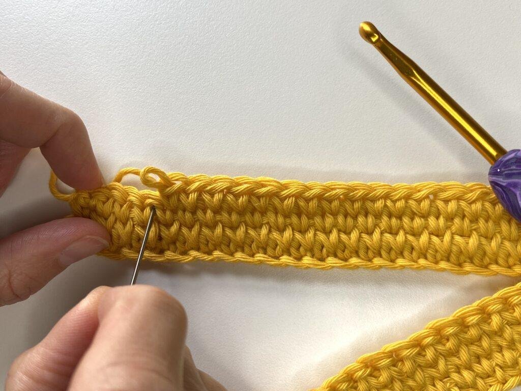 A lady pointing with a needle to the v of a crochet stitch in a piece of yellow crochet 