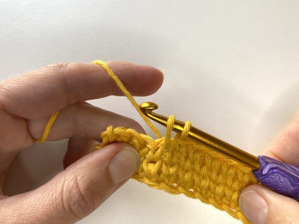 A loop is pulled through as someone continues to make a crochet stitch with yellow yarn