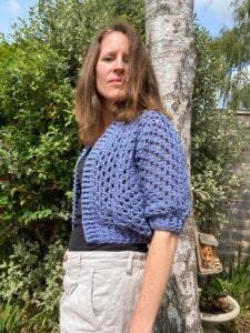 A lady stands in a garden wearing a blue crochet cardigan with a mesh effect