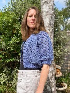 A lady stands in a garden wearing a blue crochet cardigan with a mesh effect