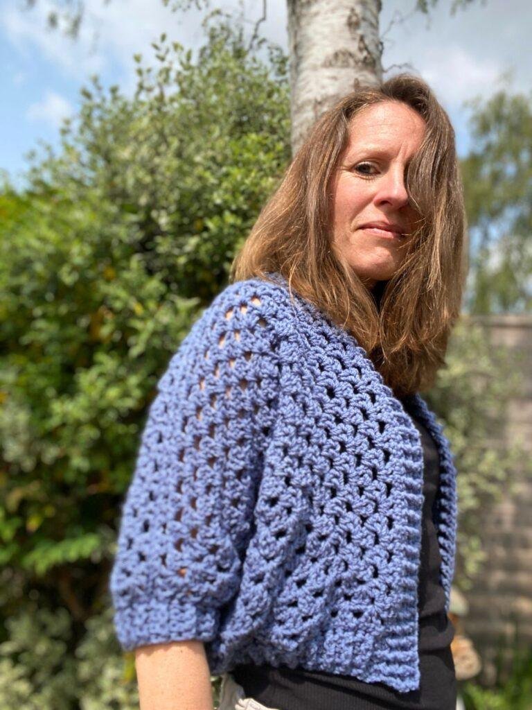 A lady stands sideways in a garden wearing a handmade crochet cardigan with three-quarter length sleeves