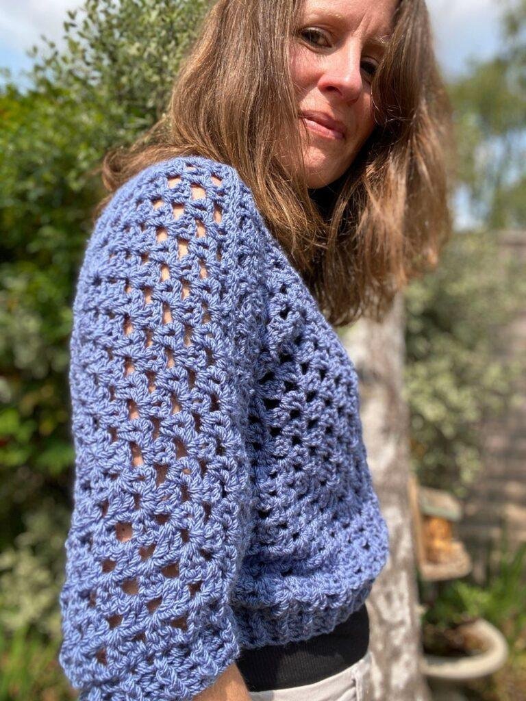 A woman stands side on in a garden looking down and wearing a crochet sweater in blue