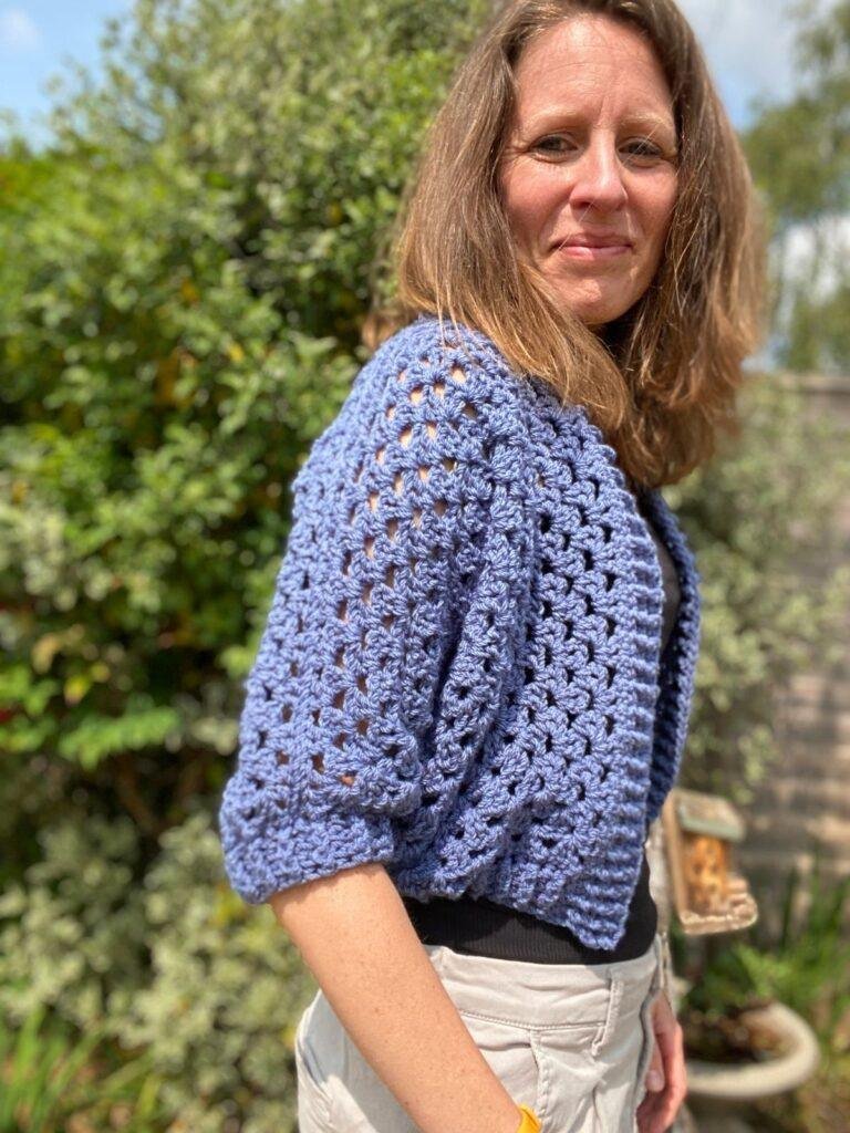 A woman standing to one side in a garden wearing cream shorts, a black tee and a blue crochet cardigan with cropped sleeves