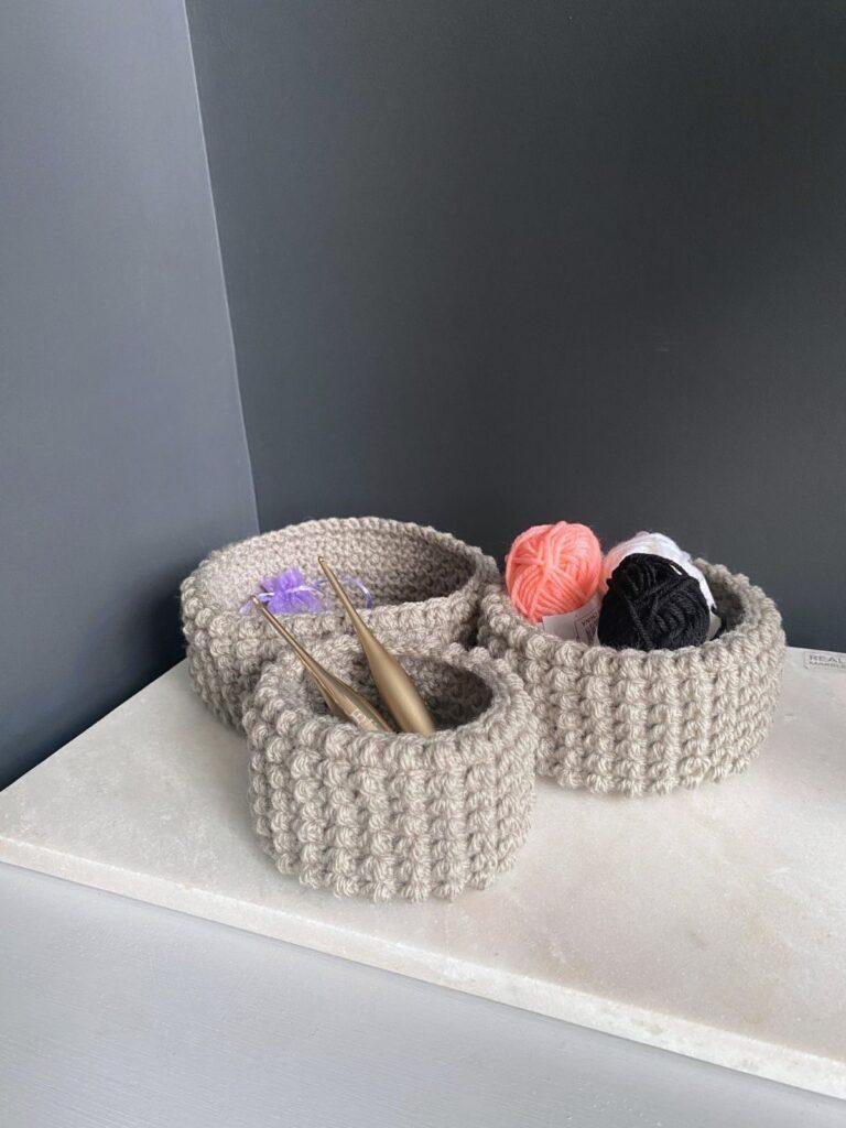 A set of 3 crochet baskets on a shelf in a corner of a grey room with crochet supplies in the baskets