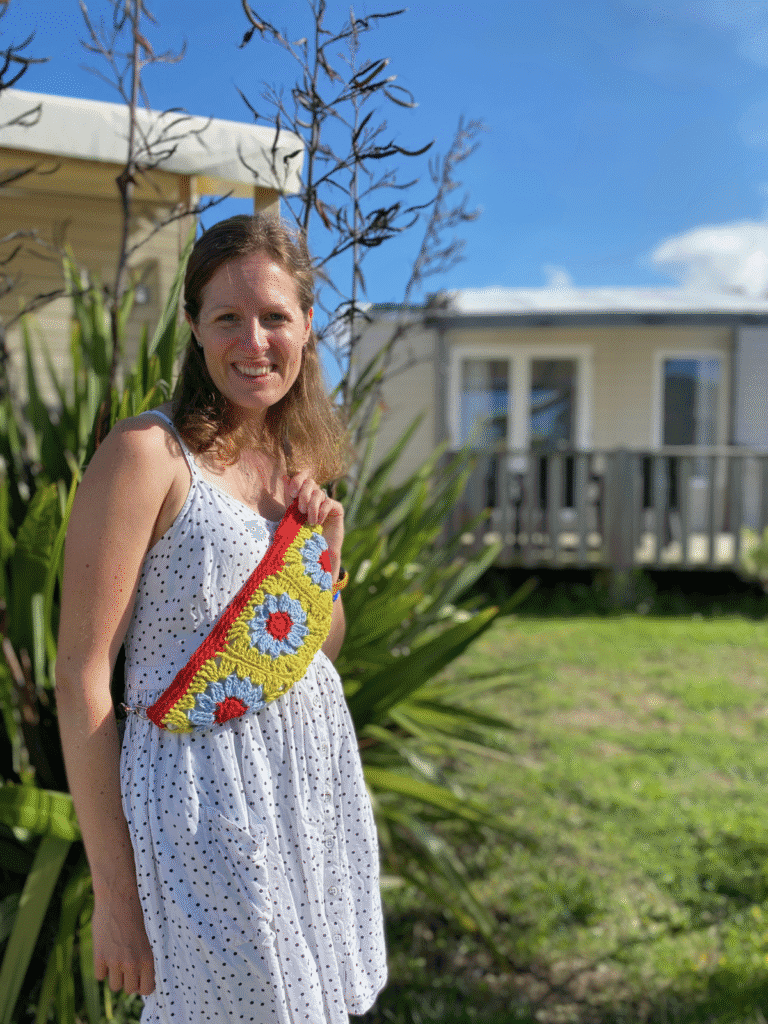 A person stands outside with a mobile home in the background. She wears a white dress and a granny square crochet bag.