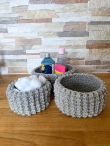 3 crochet textured baskets sit on a wooden shelf with bathroom accessories inside