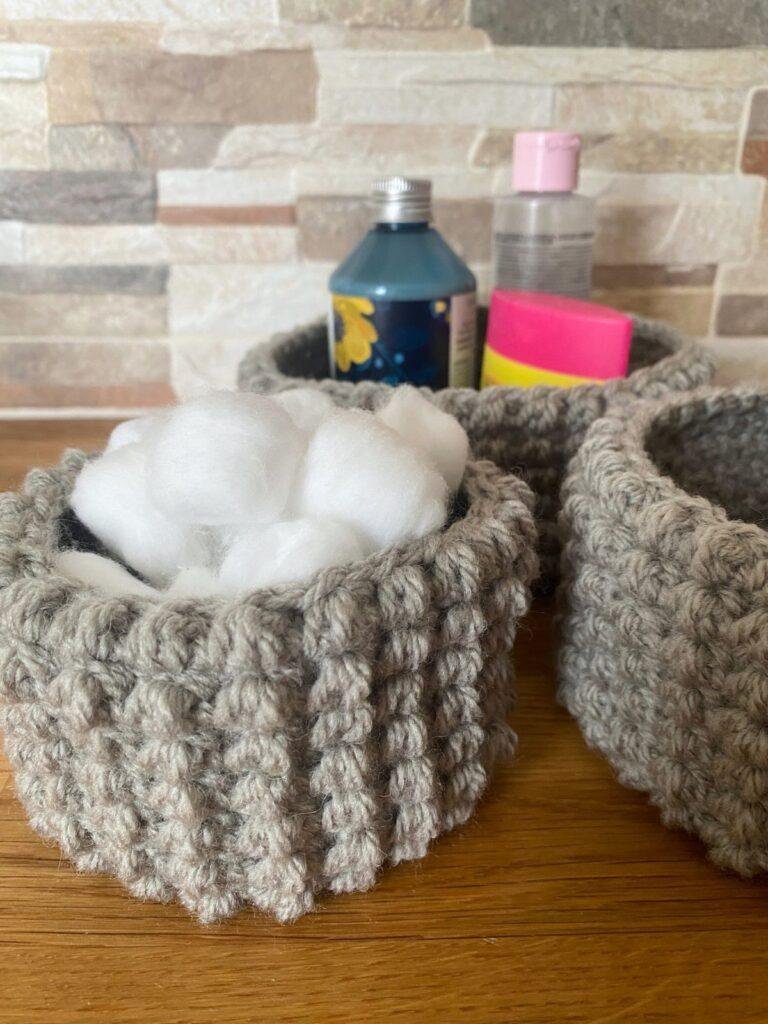 Close up of 3 grey, textured crochet baskets in a bathroom set with cotton wool, shampoo etc in the baskets