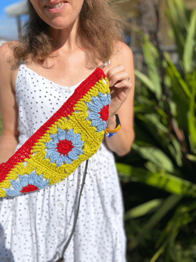 A lady stands outside by a bush wearing a white dress and holding up a trapezoid shapes crochet purse