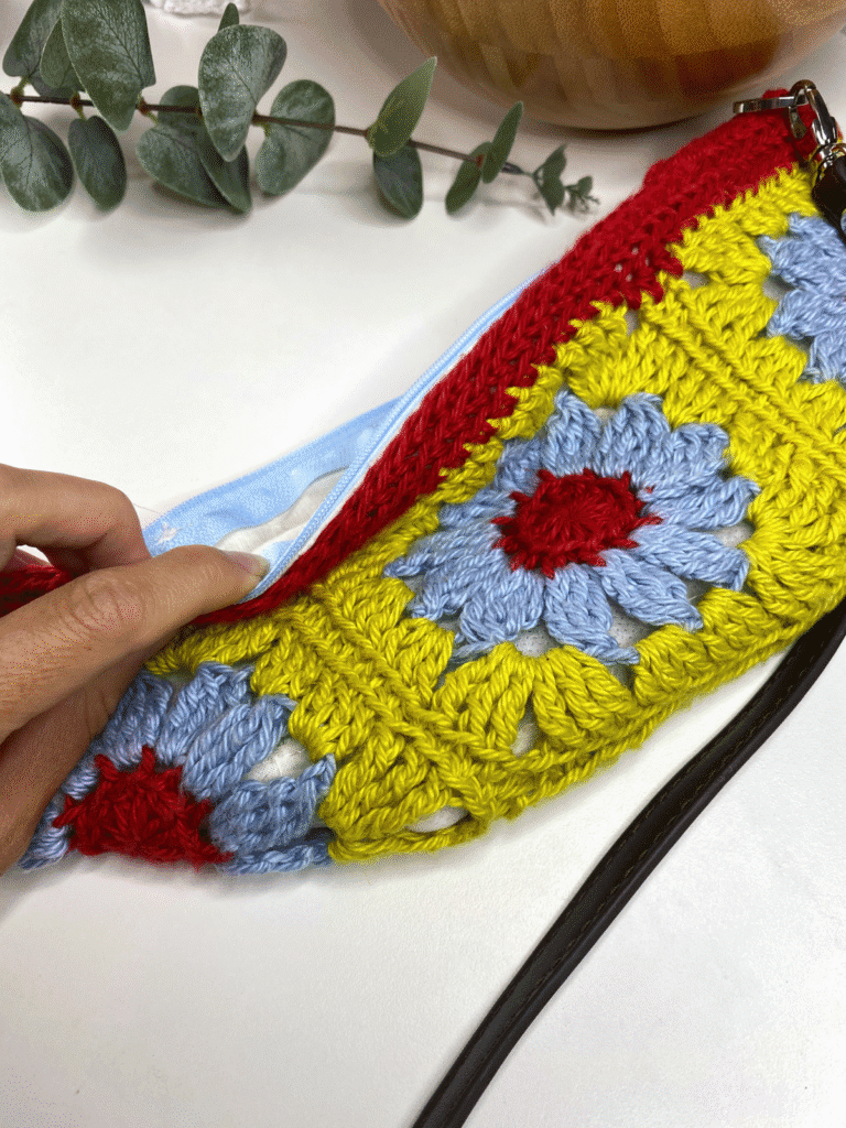 A hand is holding open a crochet purse to show to zip. The bag is green, blue and red with crochet flower motifs.